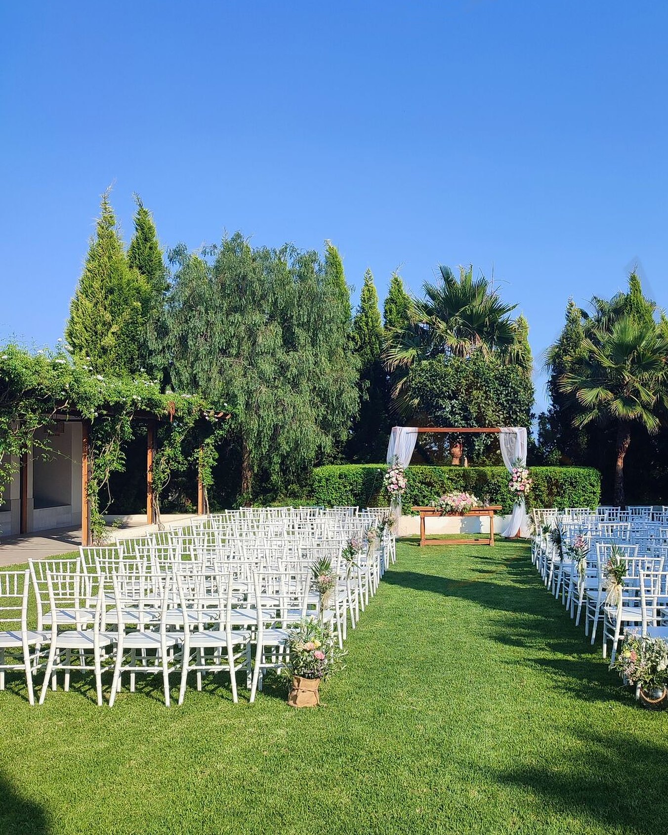 El jardín del Coso preparado para una ceremonia: sillas blancas sobre el césped y la pérgola al fondo, entre palmeras